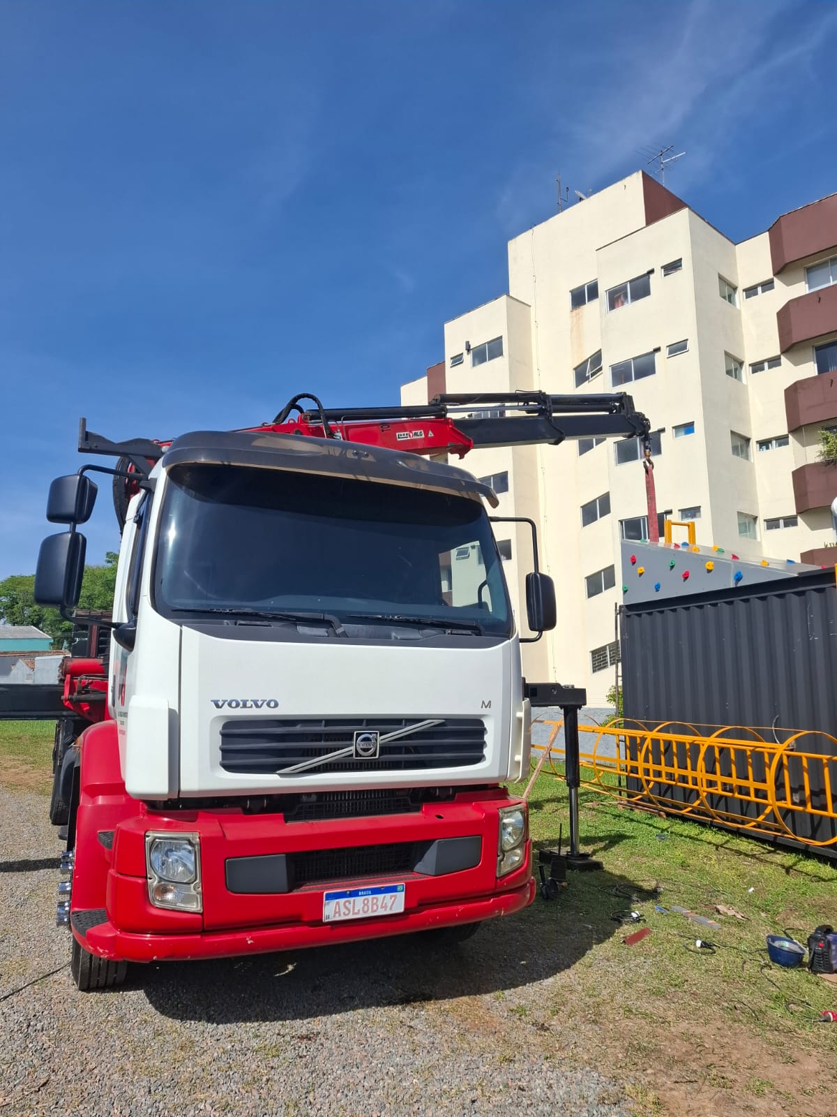 Caminhão munck Volvo VM 260 branco e vermelho da Da Veiga Muncks com a lança do guindauto estendida em operação de içamento ao lado de um edifício em Curitiba